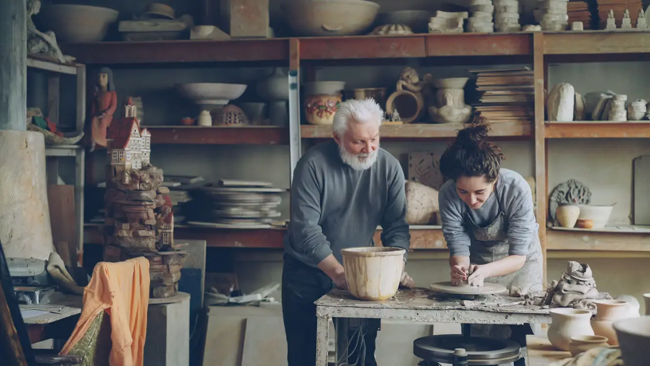 A senior craftsman guiding a younger apprentice on a pottery wheel, illustrating hands-on knowledge transfer