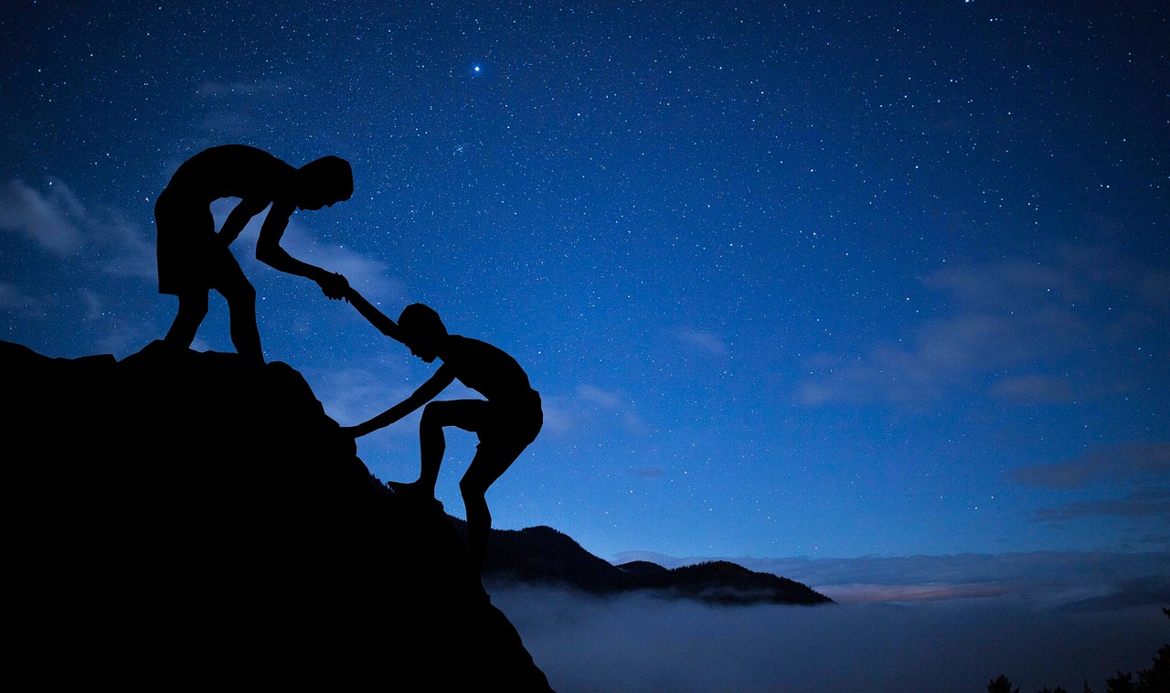 Silhouette of one person reaching down to help another climb a ridge at dusk, representing the role of a mentor in opening a path forward