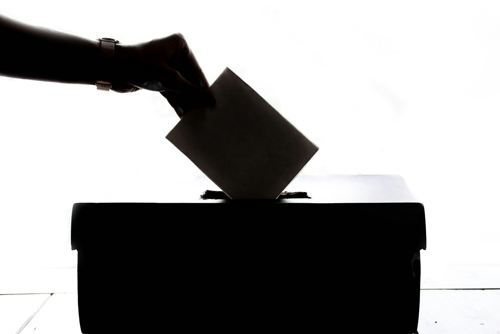 Silhouette of a hand placing a folded ballot into a ballot box against a bright white background