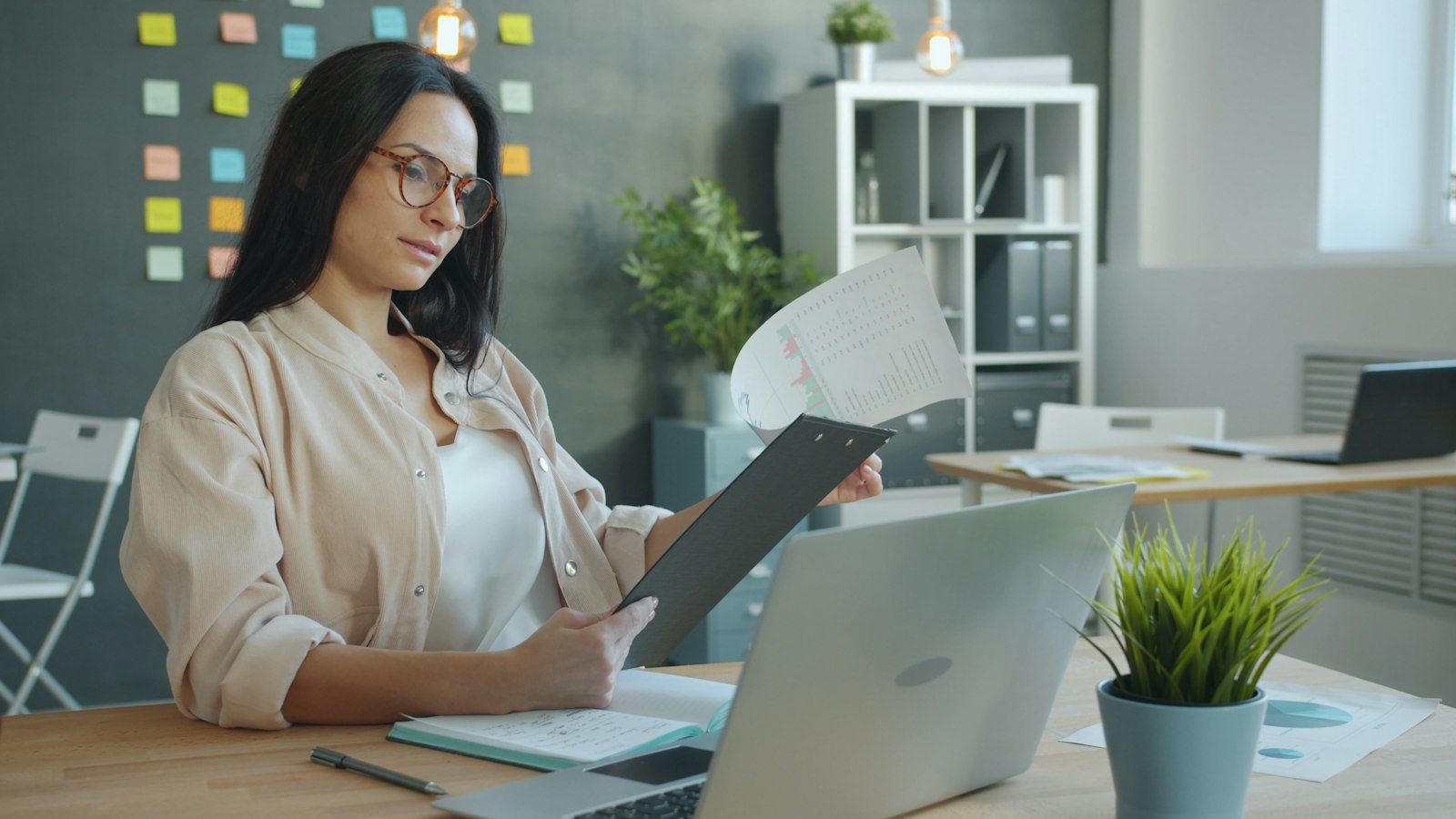 A woman in glasses reviewing a printed report on a clipboard beside her laptop, representing document-heavy knowledge work