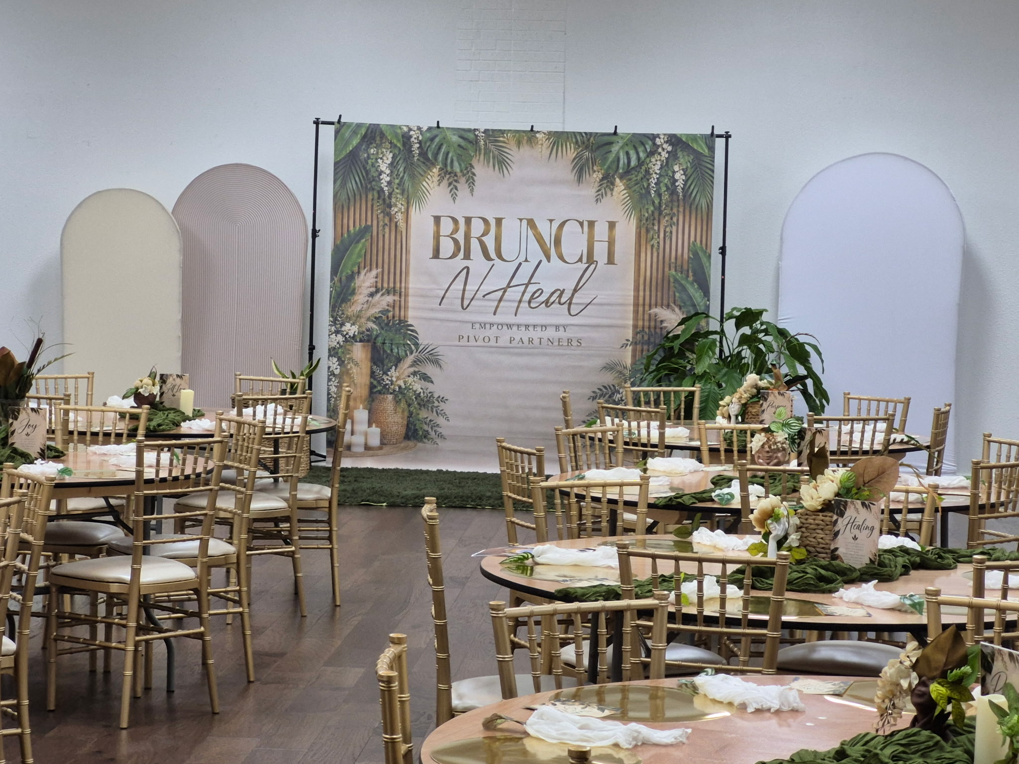 Brunch N Heal event setup with a tropical backdrop, gold chairs, and decorated round tables