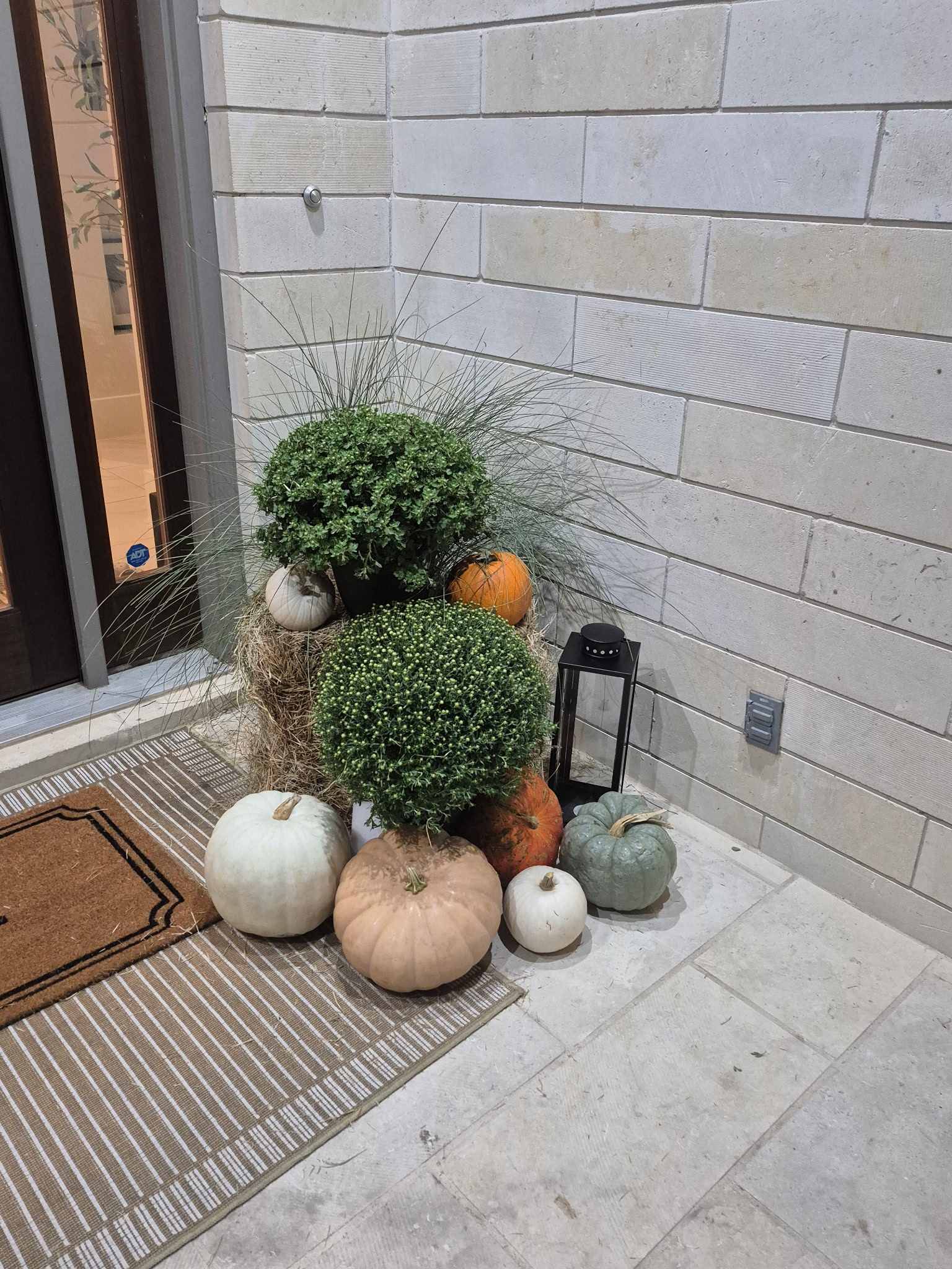 Front porch display with mums, ornamental grasses, pastel pumpkins, hay bales, and a black lantern