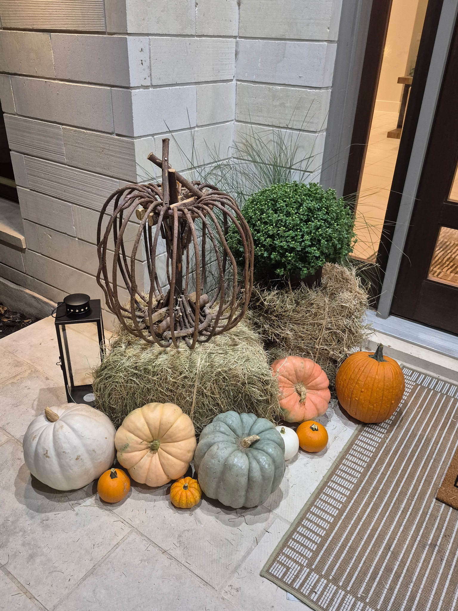 Rustic twig pumpkin on hay bales with colorful pumpkins, mums, and a lantern beside a front door
