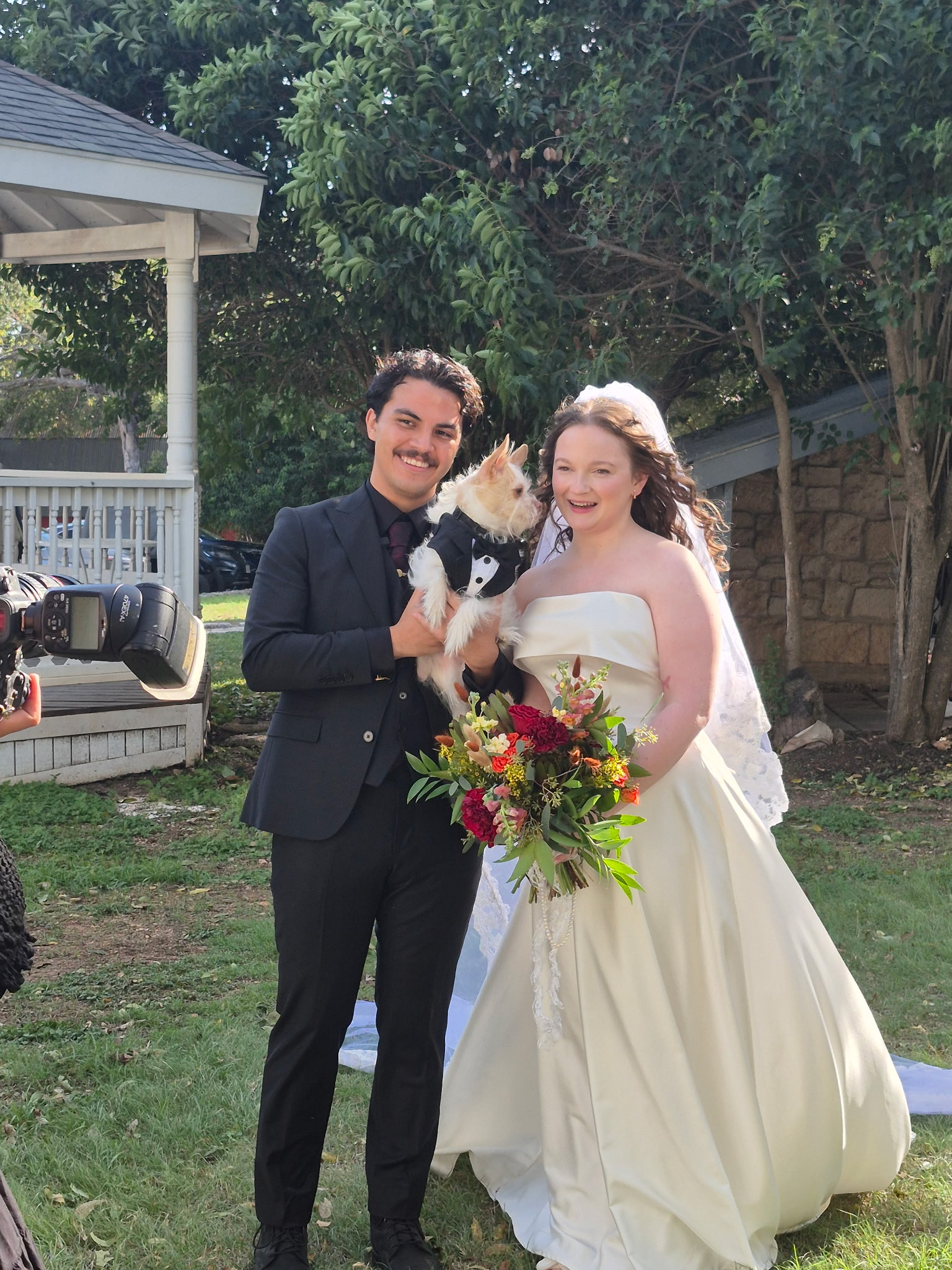 Bride and groom holding their small dog in a tuxedo outdoors on their wedding day