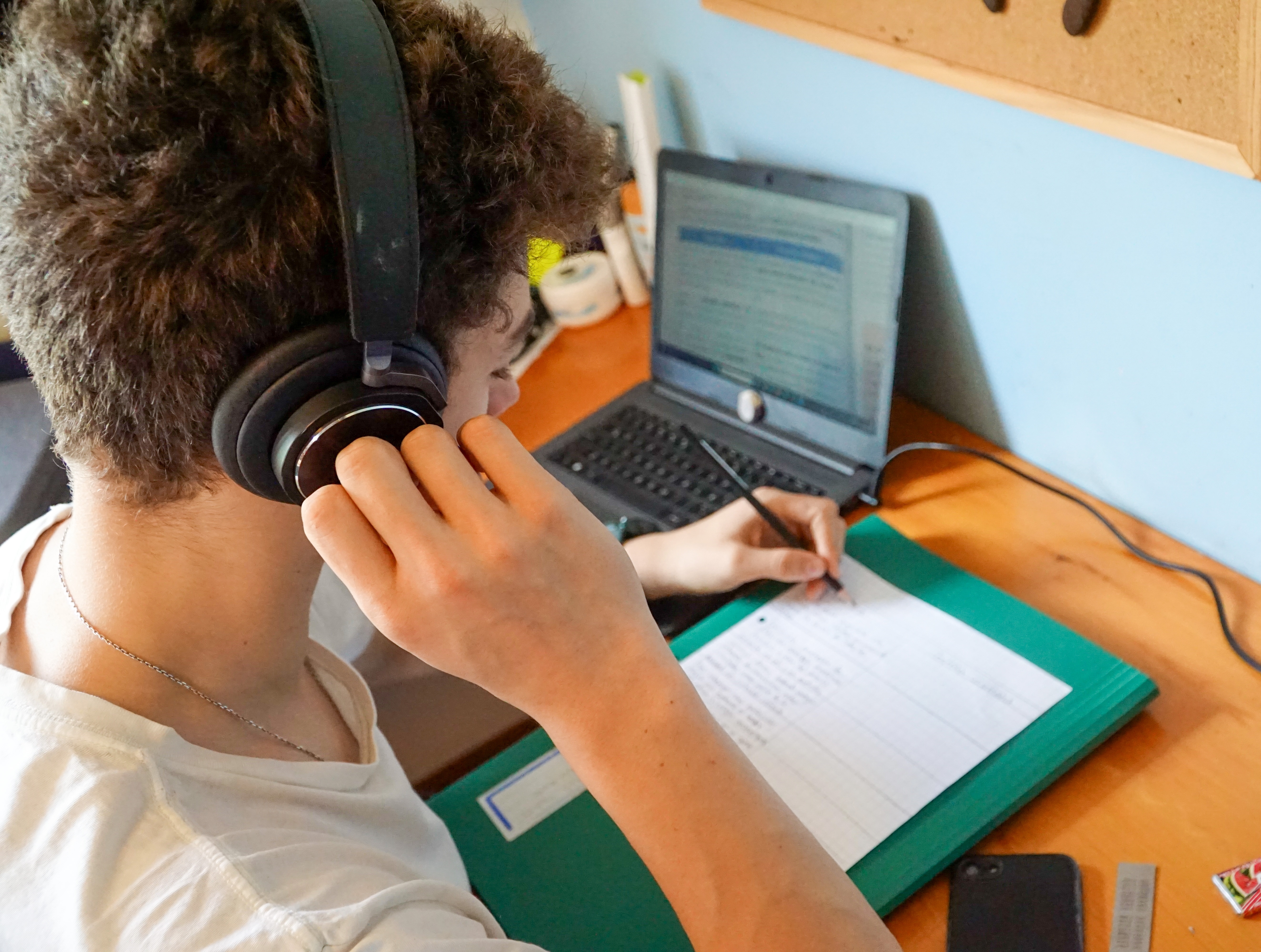 Student wearing headphones while writing in a notebook beside a laptop at a desk.