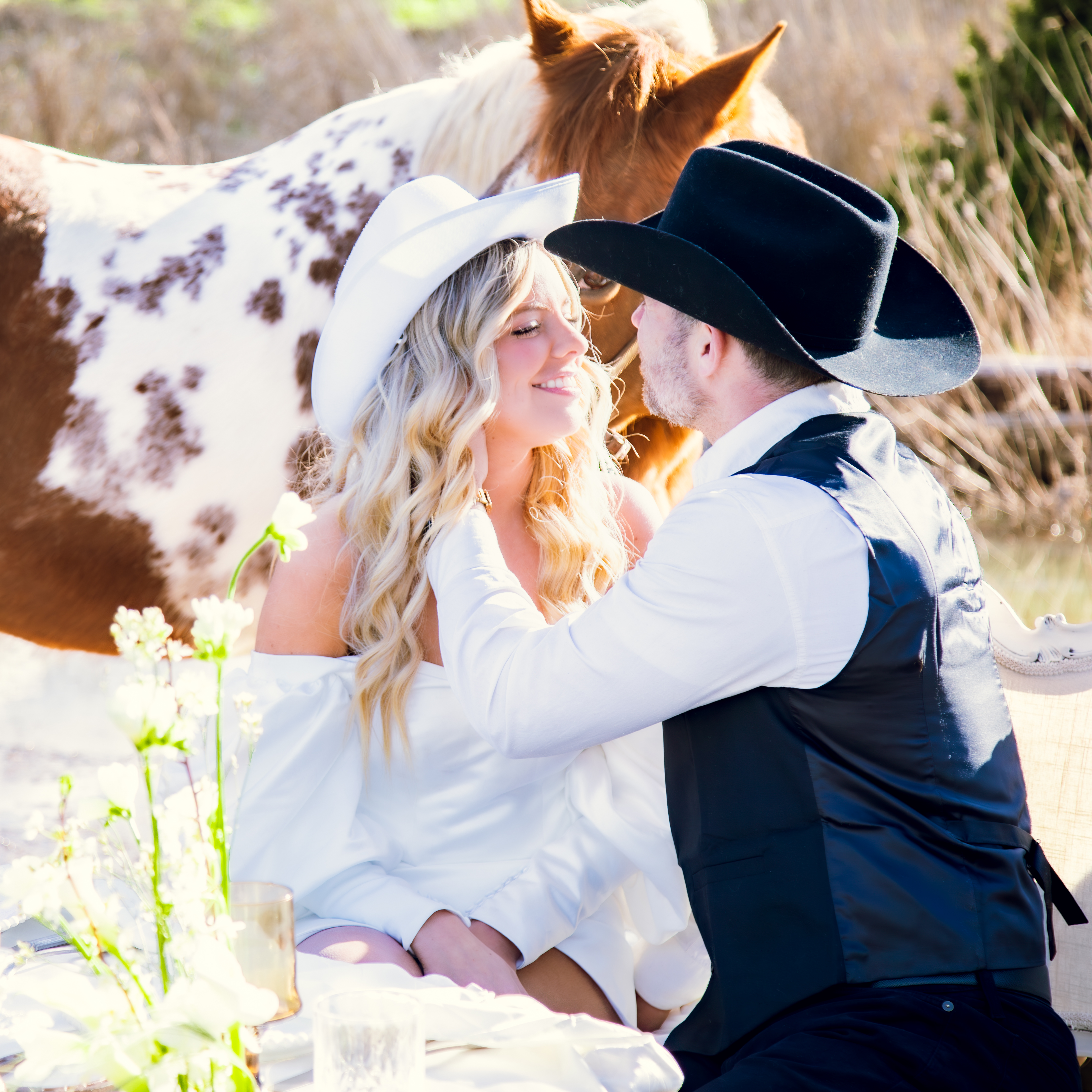 Artistic engagement portrait by Tyler Hills Photography: A happy couple in cowboy hats and a chic off-the-shoulder white dress embrace during a rustic, picnic-style styled shoot. A paint horse is visible in the soft-focus background, capturing a modern western wedding aesthetic in Salem, Oregon.