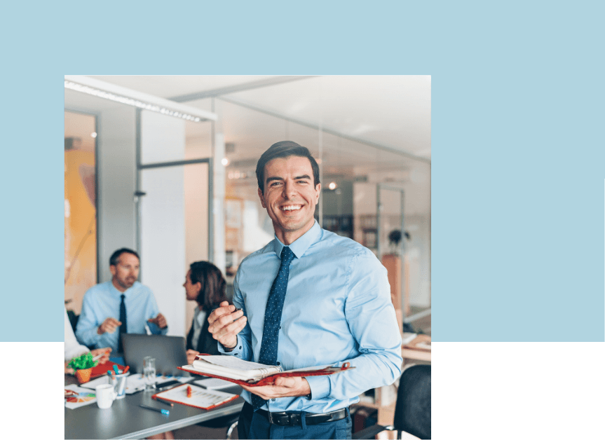 A cheerful man in an office environment, standing proudly with colleagues discussing at a table in the background, highlighting the benefits of the Emotional Footprint® Coach Certification for HR and wellness teams.