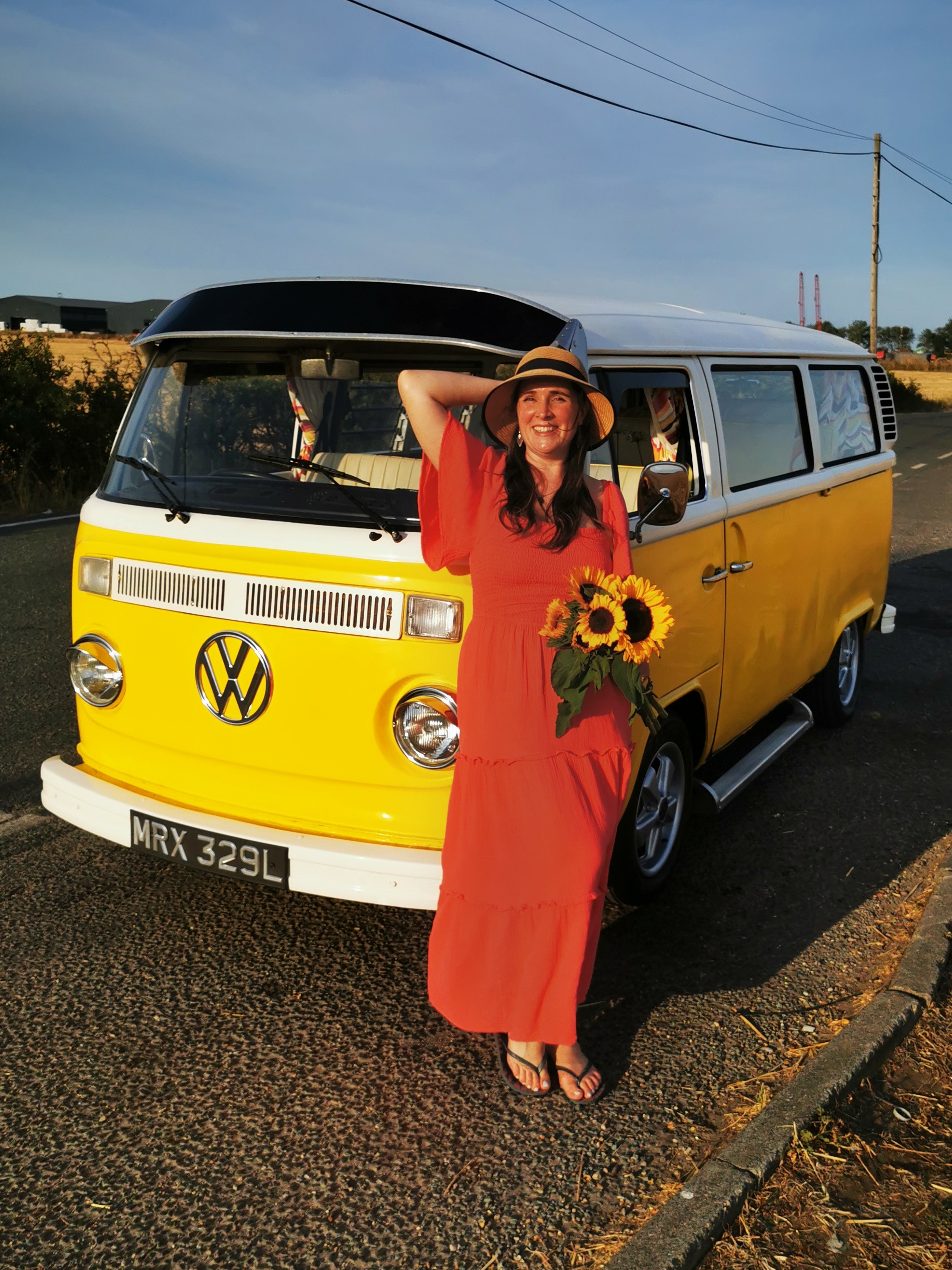A smiling woman holding sunflowers beside Marigold the vintage VW campervan