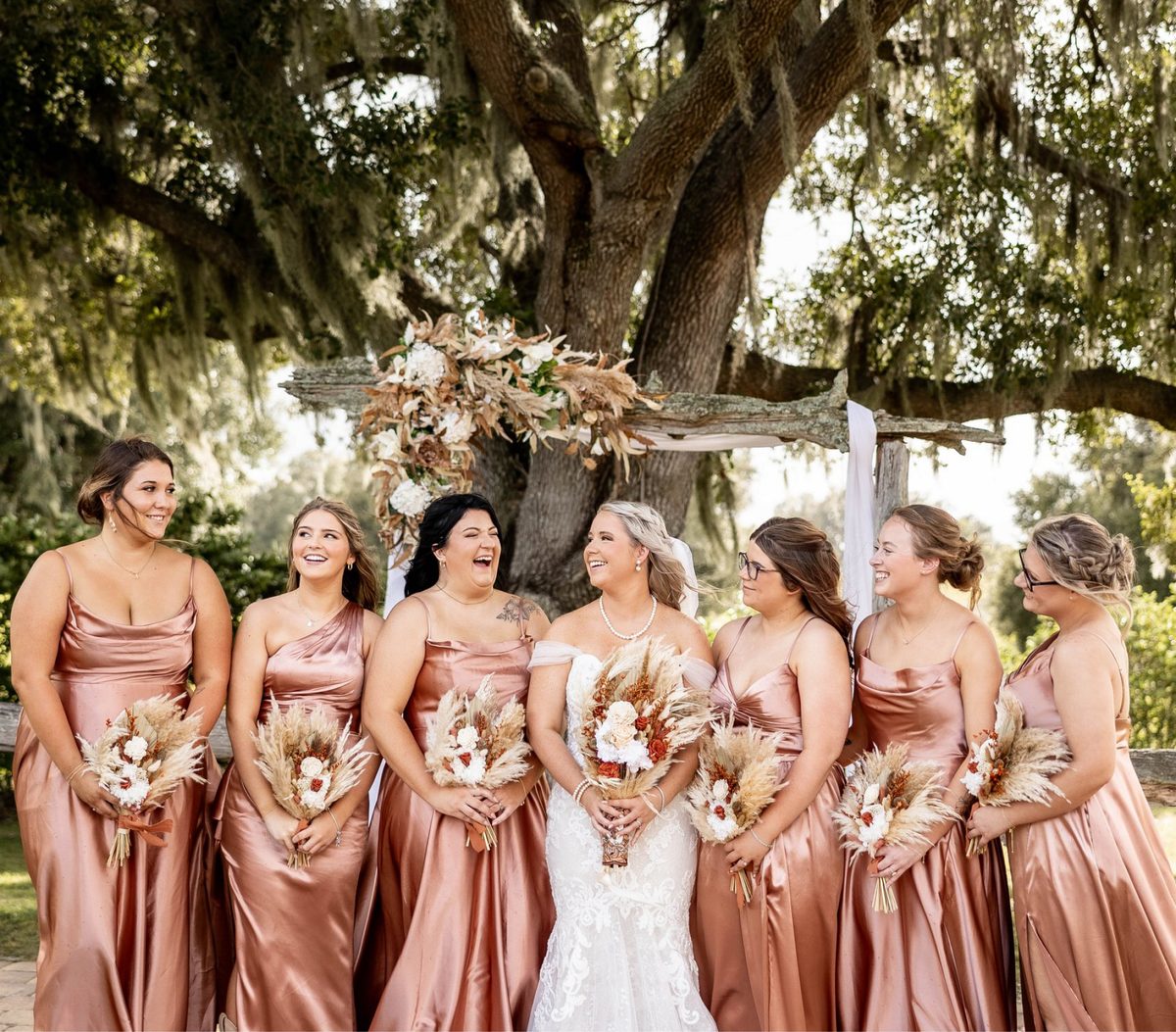 A bride laughing with her bridesmaids in rose satin dresses beneath the oaks