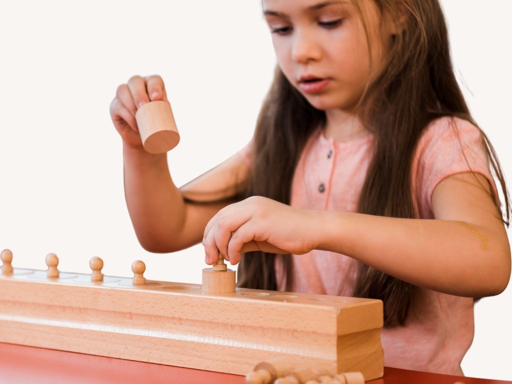 Dievča pri stolíku vkladá drevené valčeky s úchytom do Montessori bloku.
