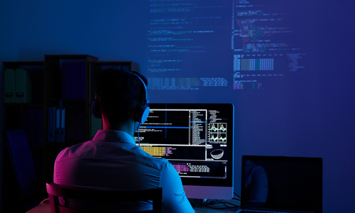 Back view of a developer wearing headphones and working on multiple screens with code and data visualizations in a dark room, representing software development and cybersecurity.