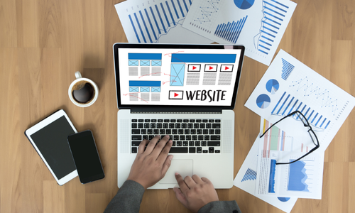 Top-down view of a desk with a person working on a laptop showing a website wireframe, surrounded by charts, graphs, tablet, smartphone, coffee, and eyeglasses, representing web development and data-driven design.