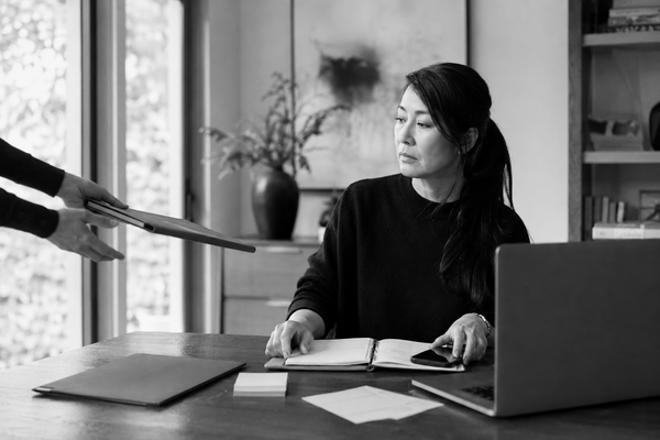 Woman at a desk managing a planner and phone while another person hands her a work folder, illustrating help that creates more mental labor