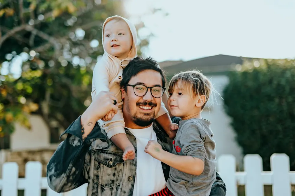 A father laughing with his two young children outside their home