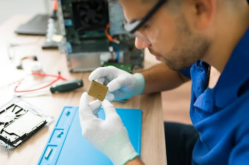 Close-up of a young technician wearing protective glasses and gloves working on a product prototype, examining a microprocessor chip.