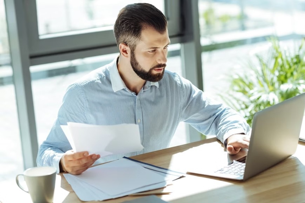 A young man sitting in an office and checking patent documentation requirement printouts while looking up applications on his laptop