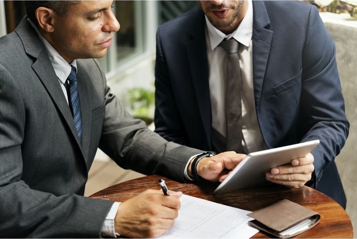 Two professionals in suits are working on an application using pen and paper and a tablet.