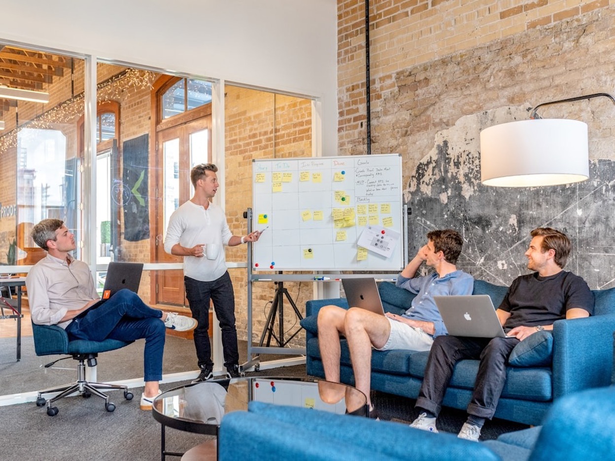 Three men are sitting, using laptops and watching a man beside a whiteboard give a presentation