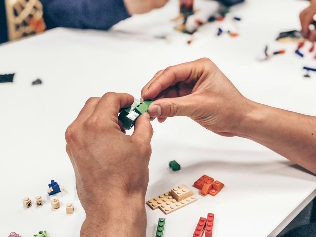 Hands assembling a toy prototype on an organized workspace with craft tools, adhesives, and labeled components laid out for step-by-step construction
