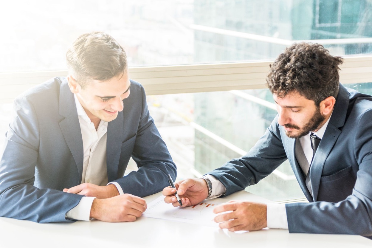 Two businessmen in suits reviewing a chart together at a desk near a bright window Two businessmen in suits reviewing a chart together at a desk near a bright window
