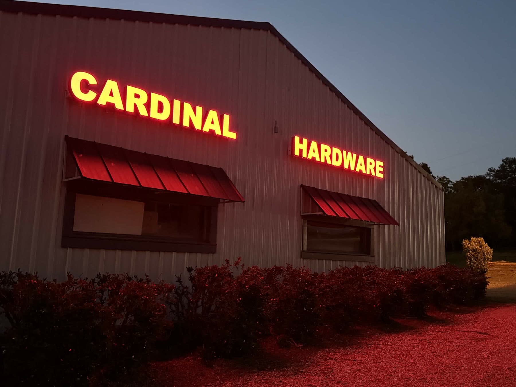 Cardinal Hardware storefront at dusk — wide view of the lit-up red CARDINAL HARDWARE letters glowing on the metal building at 104 S Laurel Street, Summit, MS