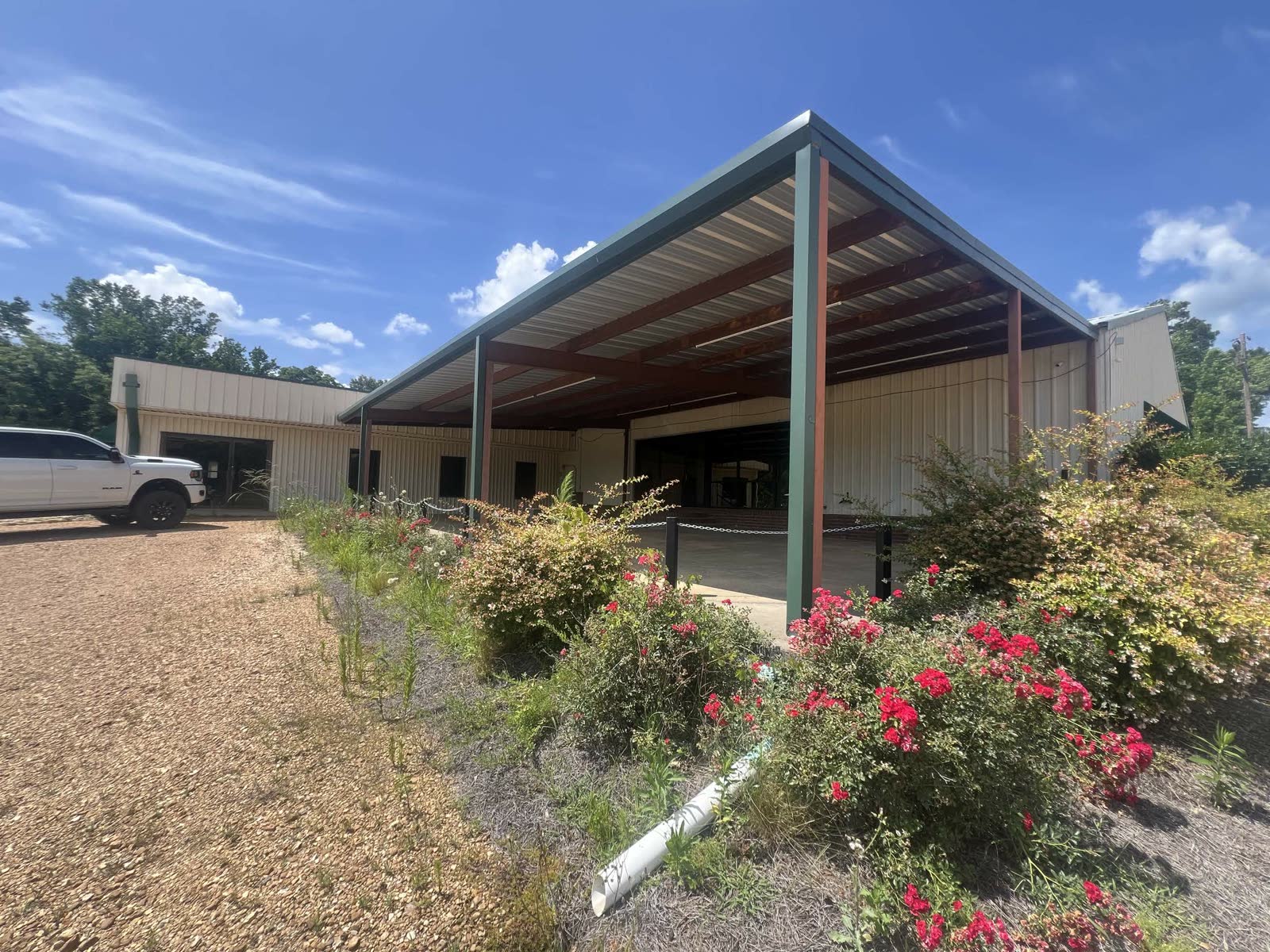 Exterior in summer — bright blue sky, flowers blooming in front, the open porch and gravel parking ready, still no signage
