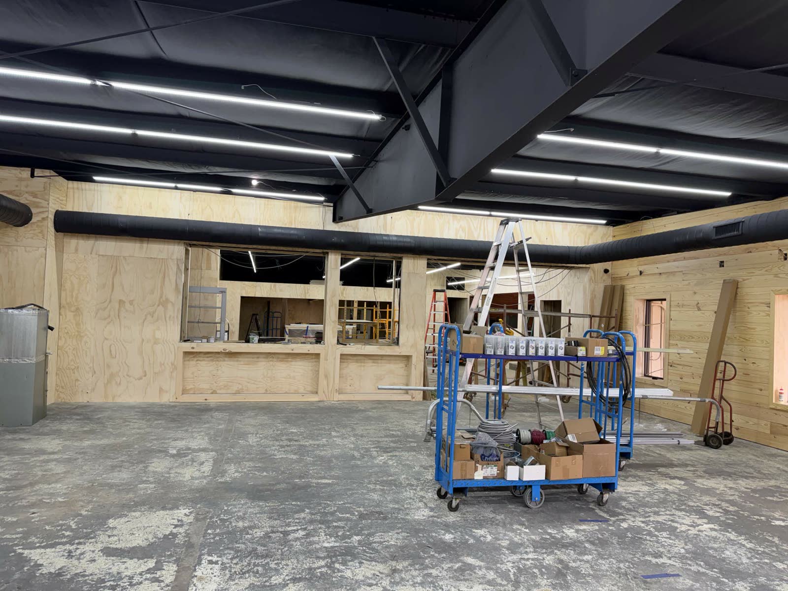 Mid-construction — black ceiling, exposed black ductwork being run, plywood walls in place with cutouts for the future service counter
