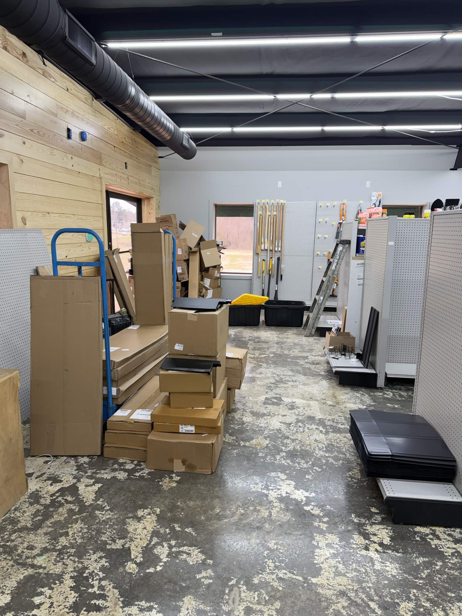 Stocking phase — stacks of cardboard boxes, white pegboard shelving units, brooms and yard tools propped against the wall