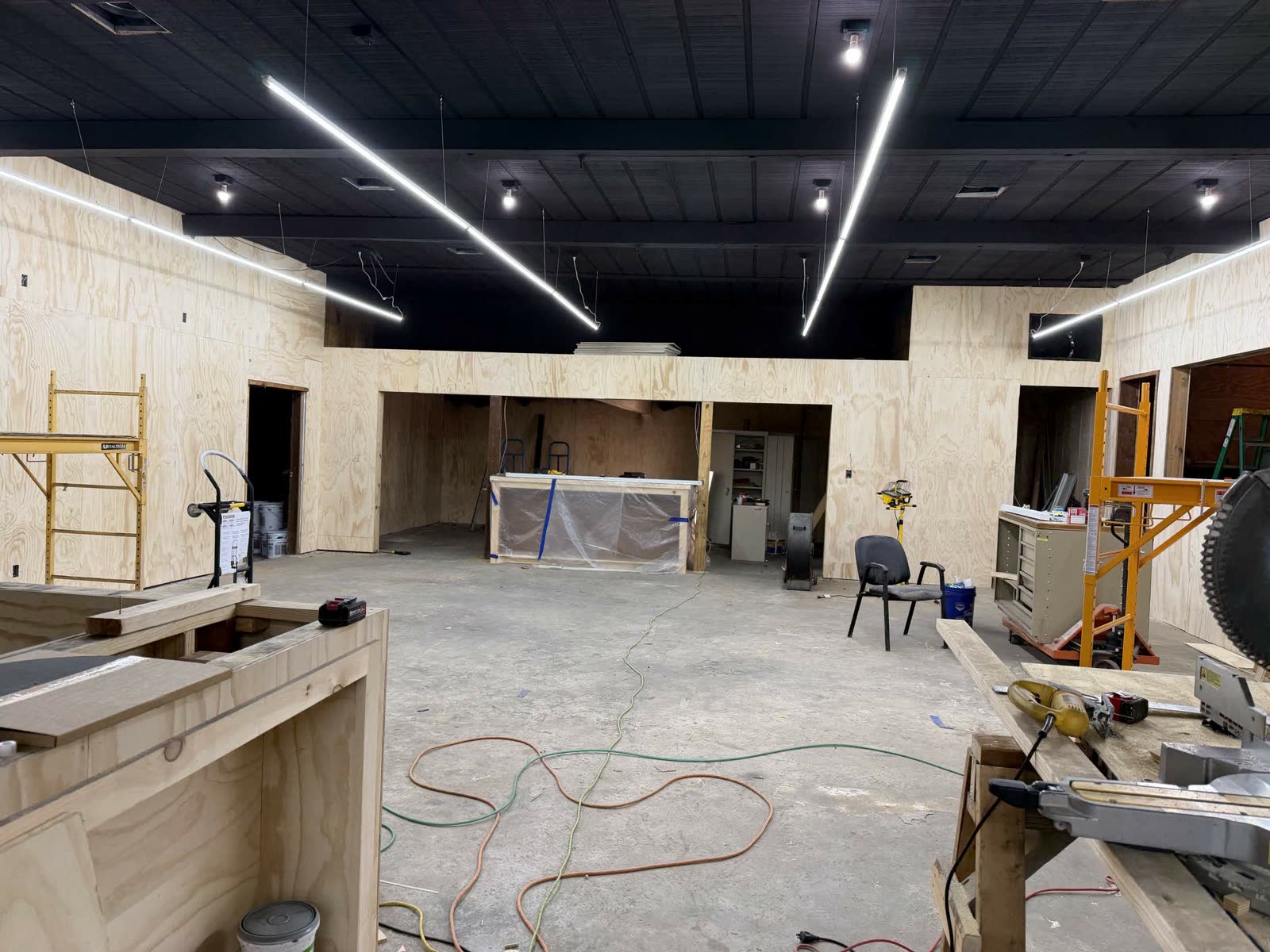 Mid-construction — fresh plywood walls throughout, modern LED strip lighting, polished concrete floor, table saw set up in the middle of the room