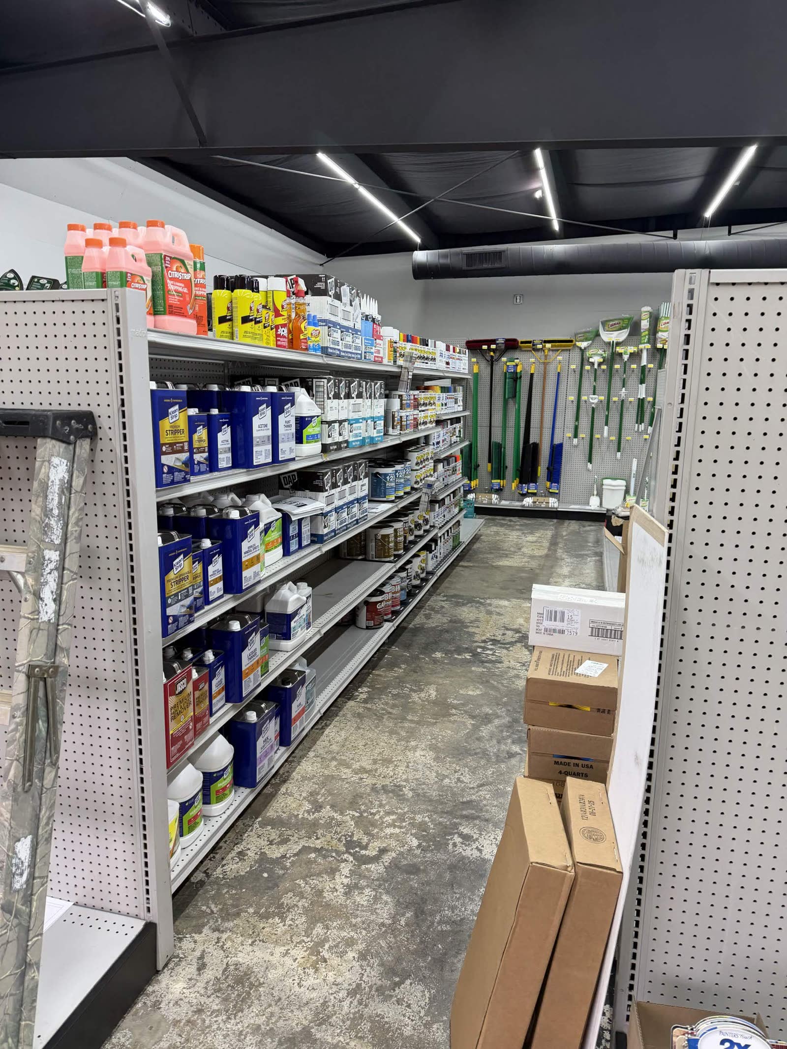 Stocking phase — store aisles filling up, lawn tools (rakes and brooms) hanging on the wall in the green-handled section
