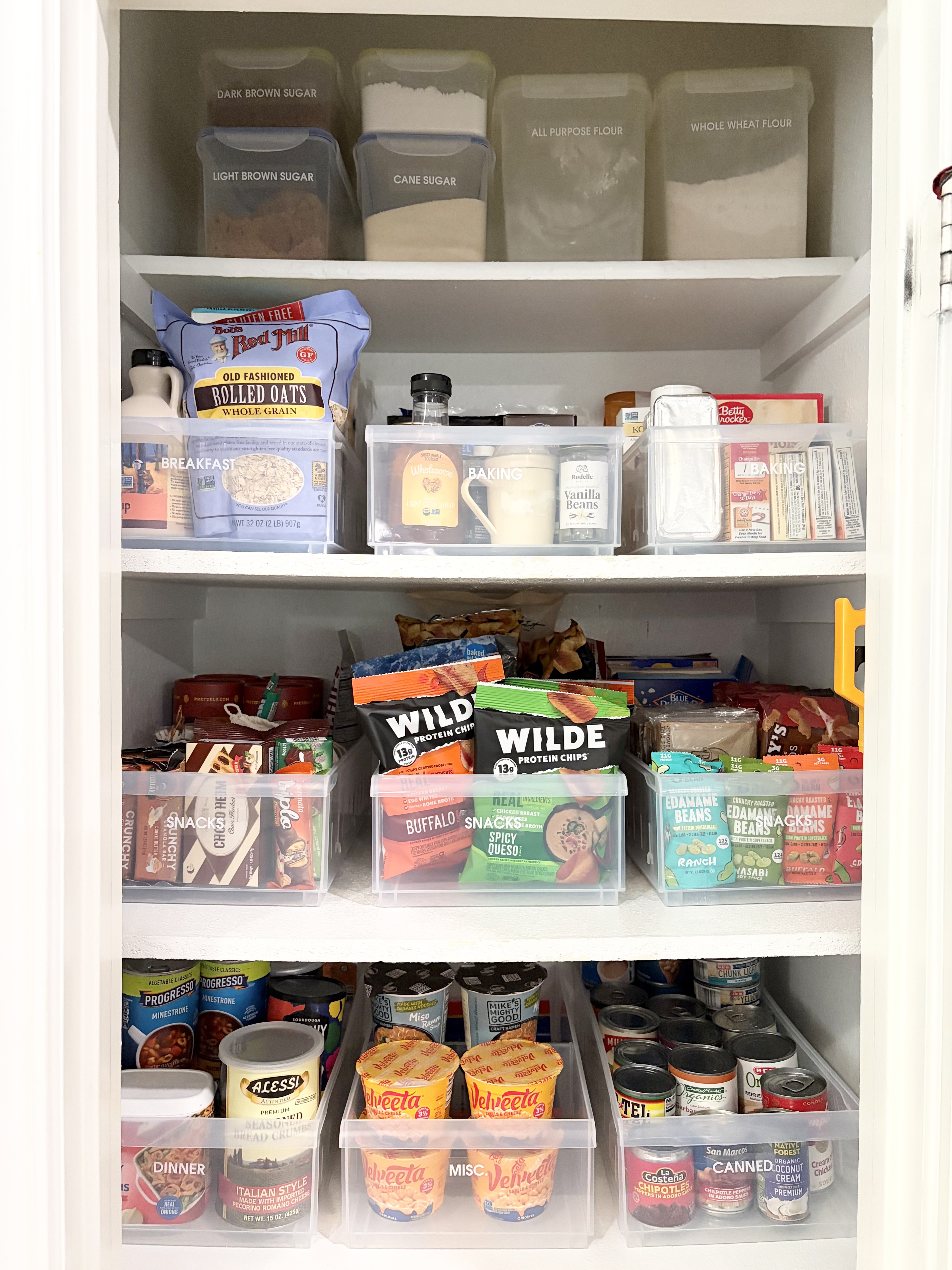 Narrow, deep builder-grade pantry closet organized with long clear pull-out bins and labeled canisters