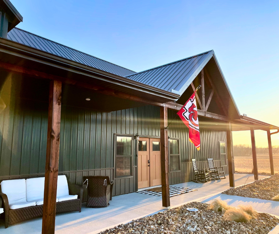 Modern farmhouse exterior with a metal roof, welcoming porch featuring rocking chairs, white sofa, and a Kansas City Chiefs flag, set against a sunset backdrop.