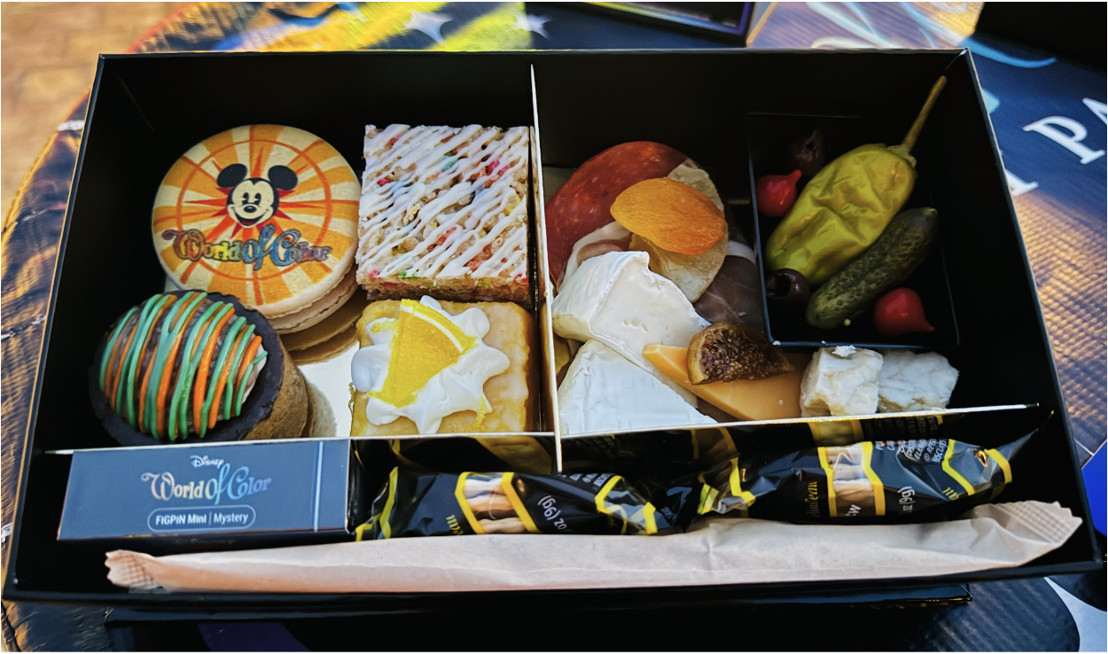 World of Color Dessert Party food box at Disneyland Resort featuring a Mickey Mouse macaroon, colorful drizzled brownie, lemon tart, rice krispy, artisan cheese and charcuterie board, pickled vegetables, and a World of Color FiGPiN Mini Mystery collectible pin]