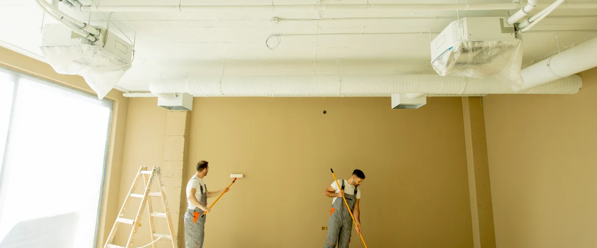 Two professional painters applying beige paint to an interior wall in a commercial space. Two professional painters applying beige paint to an interior wall in a commercial space.