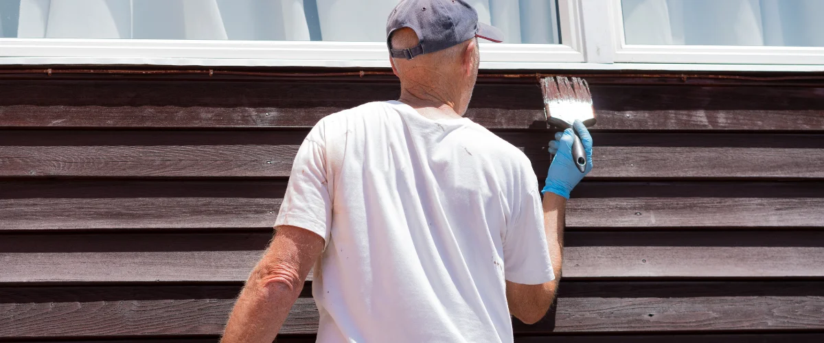 Man painting wooden house siding with brush for exterior maintenance.