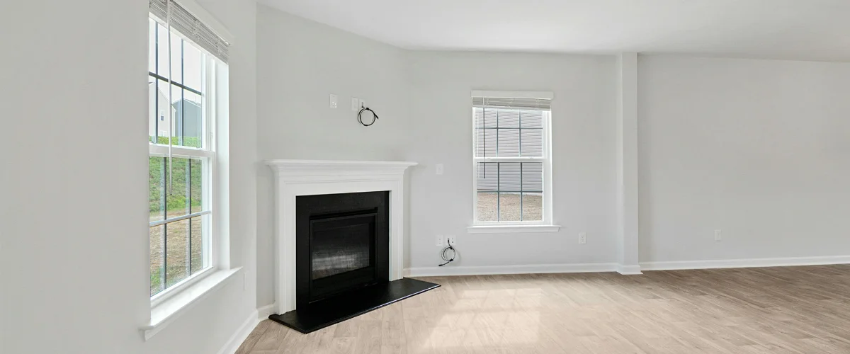 An empty living room with light hardwood flooring, light gray walls, a black fireplace, and two large windows.