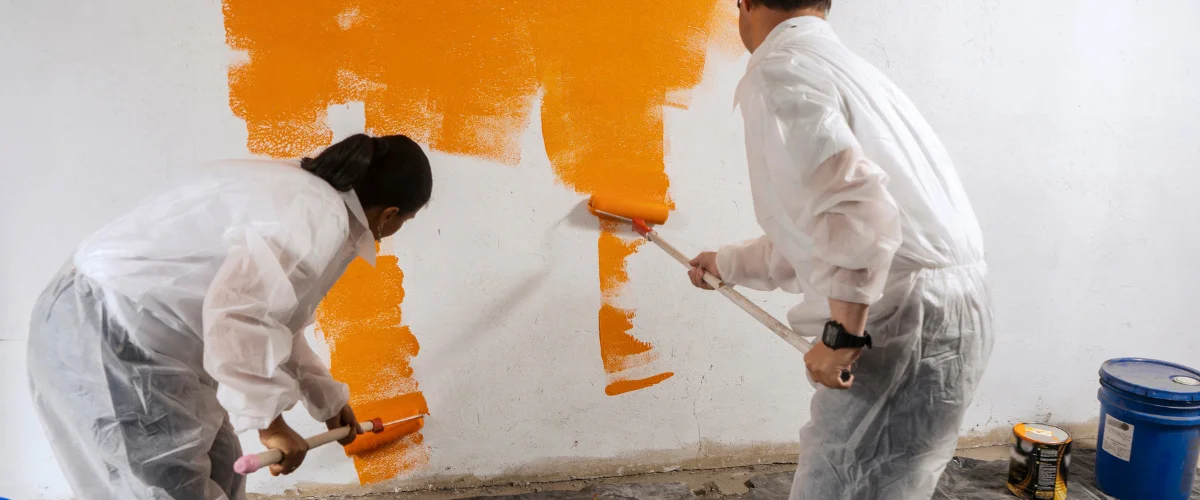 Two individuals in white protective suits painting a textured white wall in vibrant orange with paint rollers.