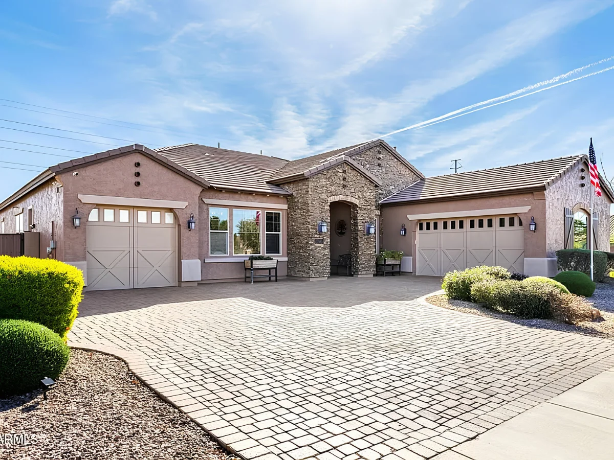 A tan stucco and stone house with a curved brick paver driveway under a bright blue sky.