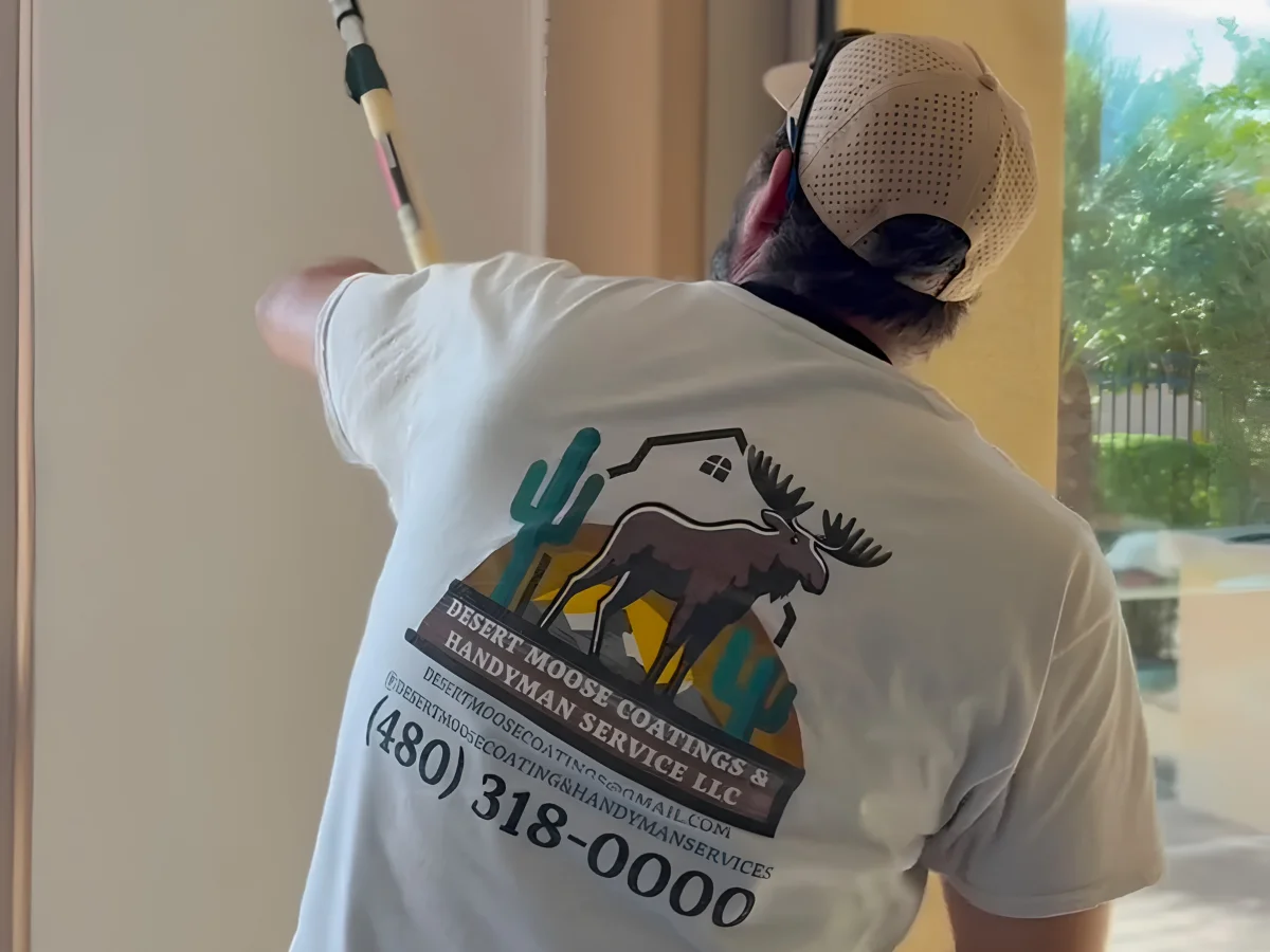 A person from behind wearing a white t-shirt with a Desert Moose Coatings & Handyman Service logo, painting a wall.