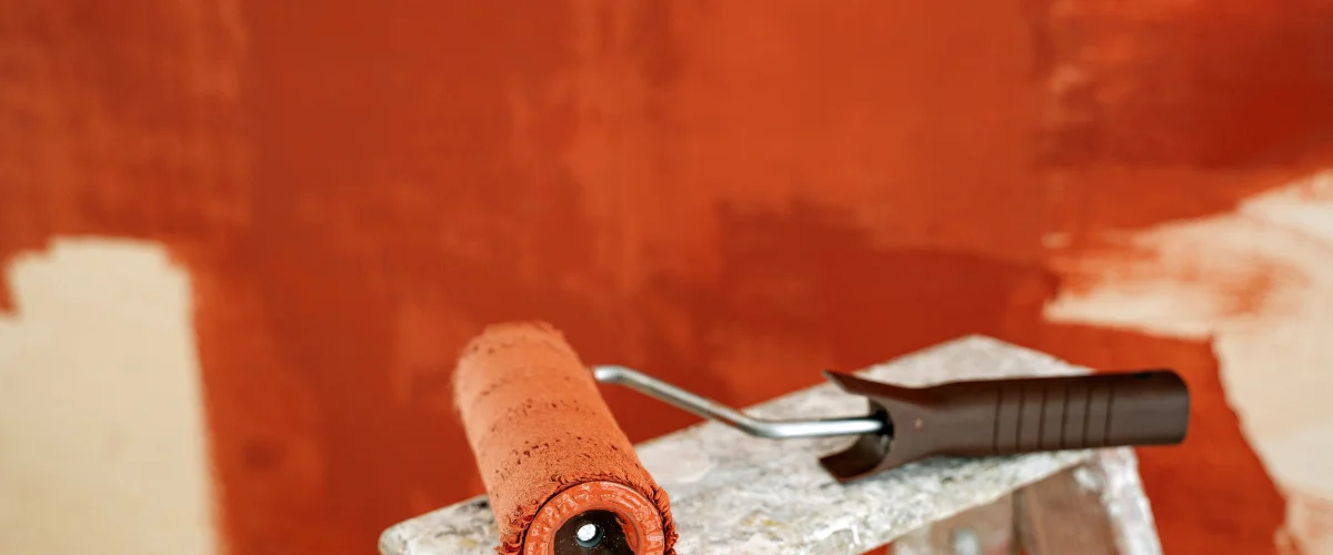 A paint roller with rust-colored paint rests on a step ladder, with a partially painted orange-red wall behind it.