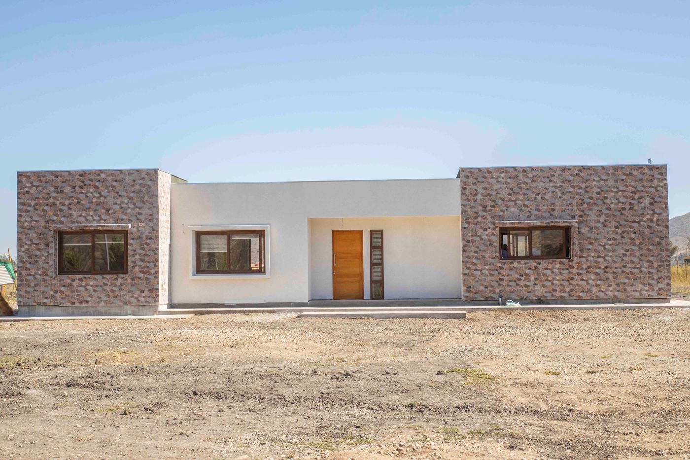 Fachada de casa nueva construida por Fortafe, con muros revestidos en piedra, puerta de madera y ventanas color nogal