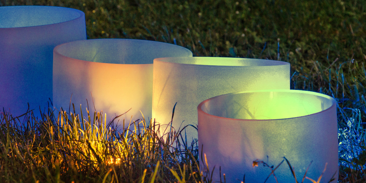 Crystal singing bowls outdoors on grass with coloured glow, used for relaxing sound bath sessions