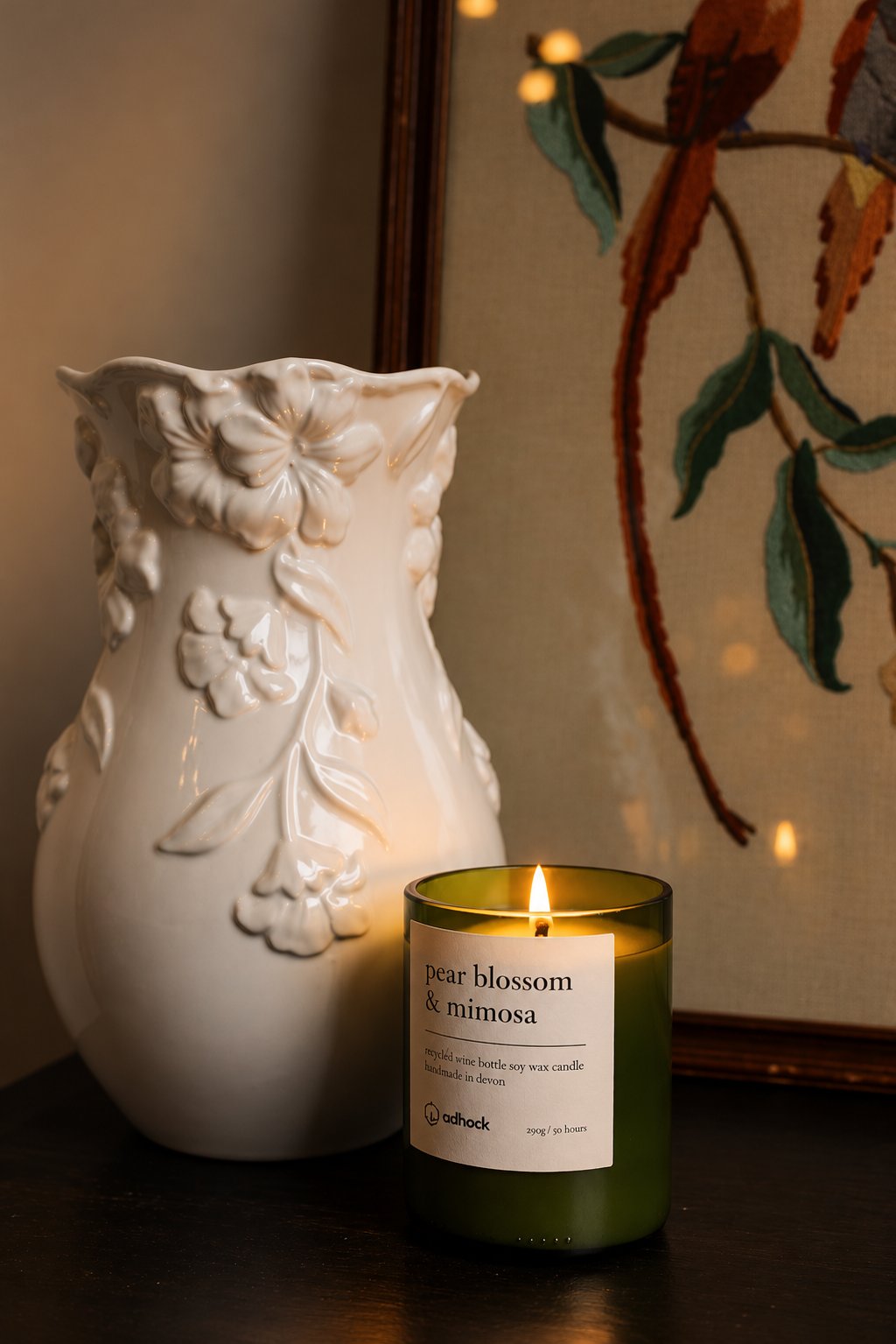 A lit pear blossom and mimosa candle beside a white floral vase