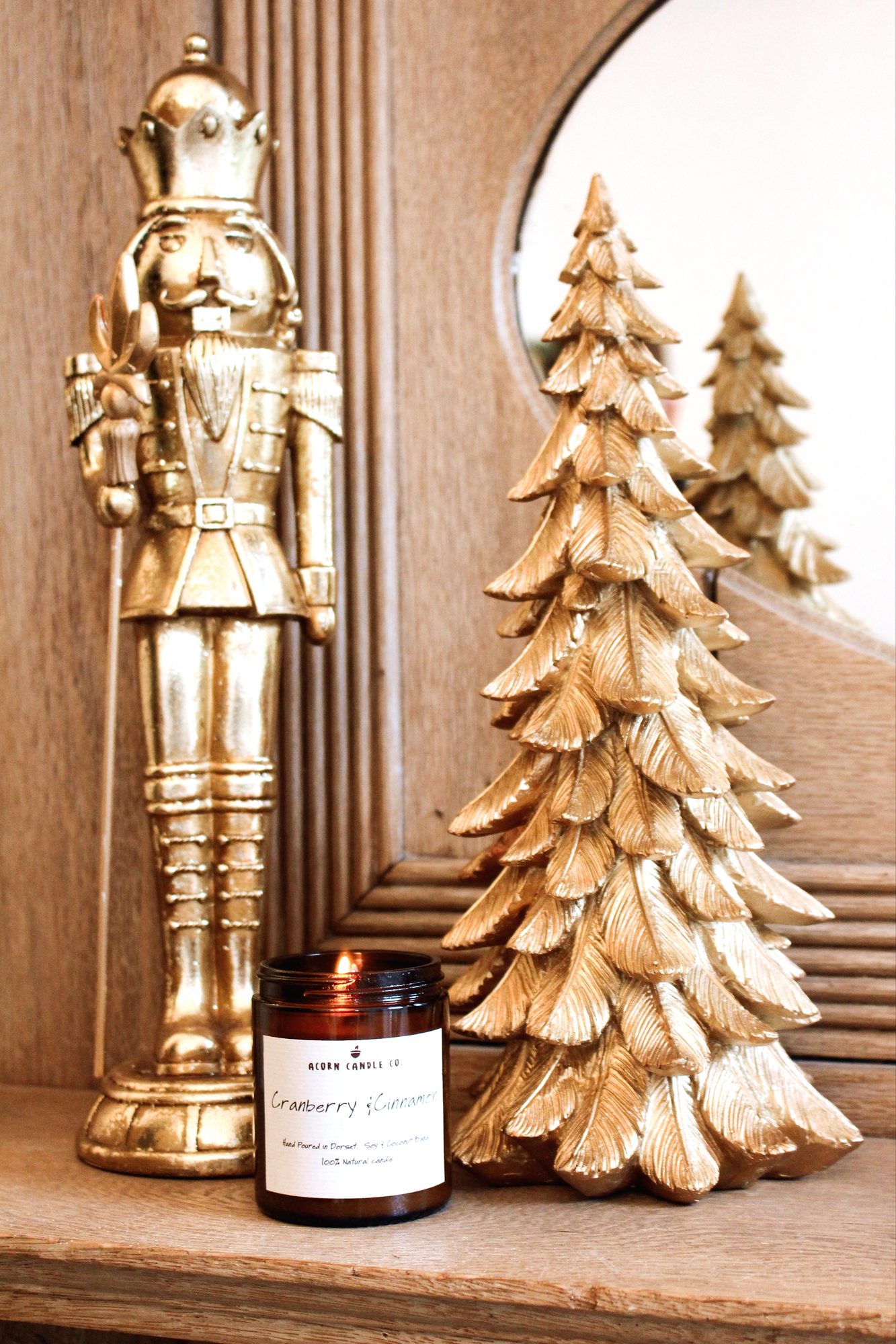 A lit Cranberry and Cinnamon candle beside a gold nutcracker and gold tree