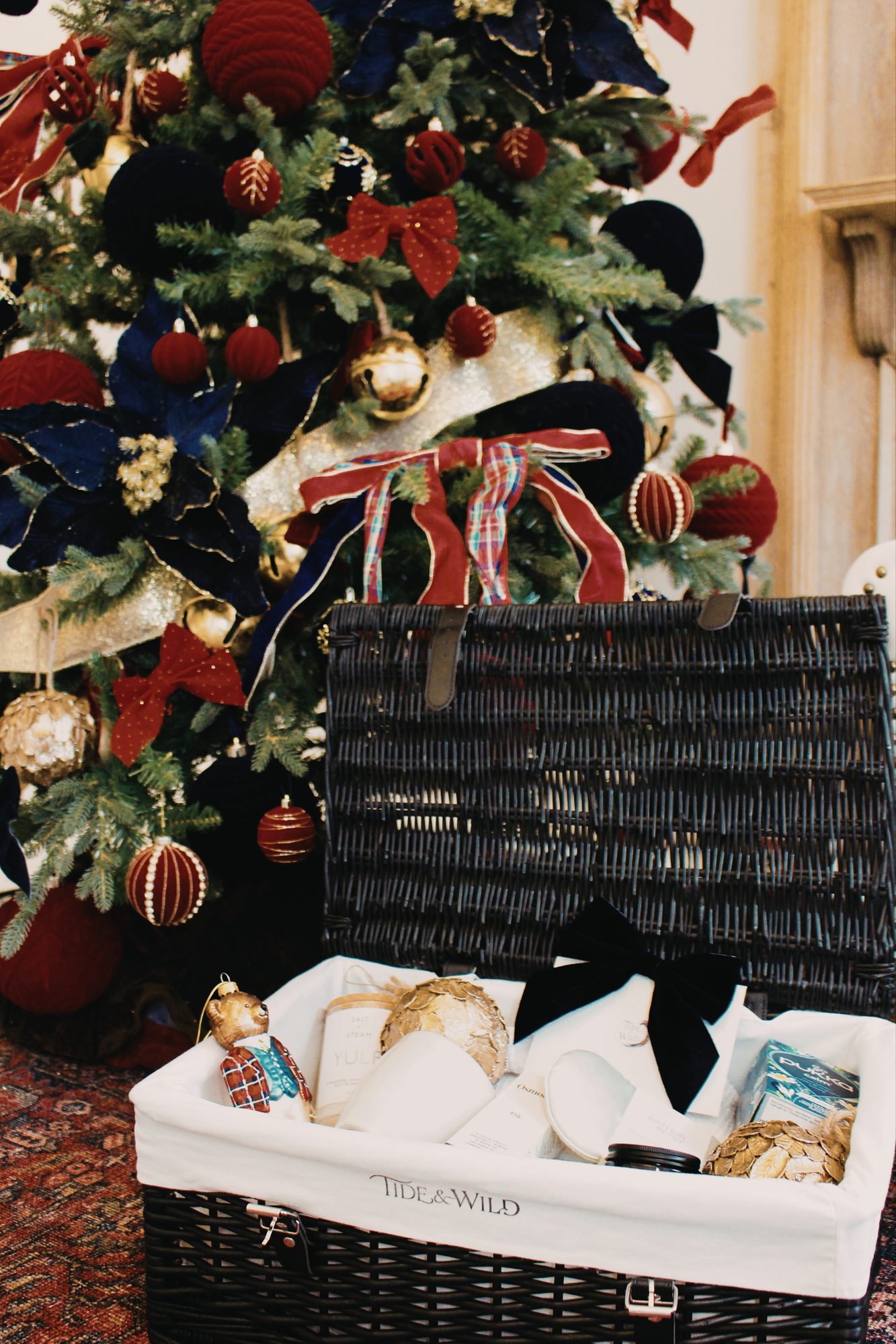 The Yule Tide Slumber hamper open in front of a Christmas tree, with a black velvet bow and gold baubles