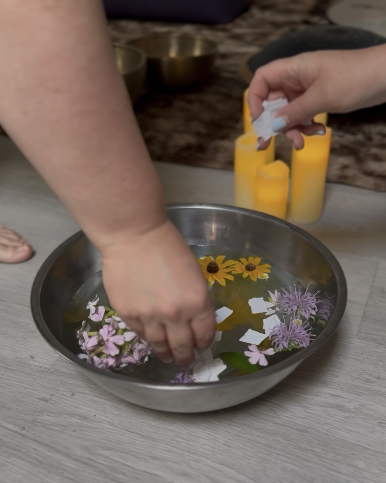 Two hands sprinkling ripped up pieces of paper into a metal bowl of water with flowers floating in it