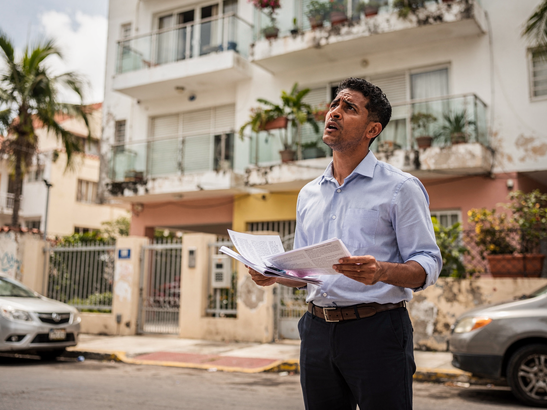 Hombre dominicano confundido frente a edificio de apartamentos con documentos en mano