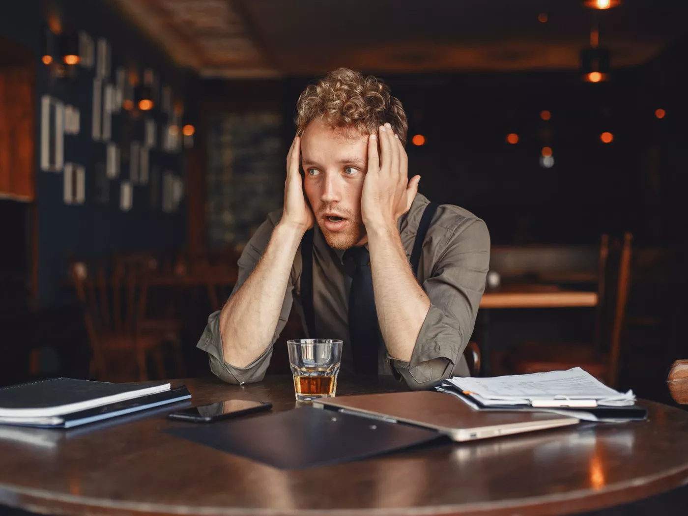 A stressed man with hands on head, surrounded by work materials, sits in a dimly lit restaurant.