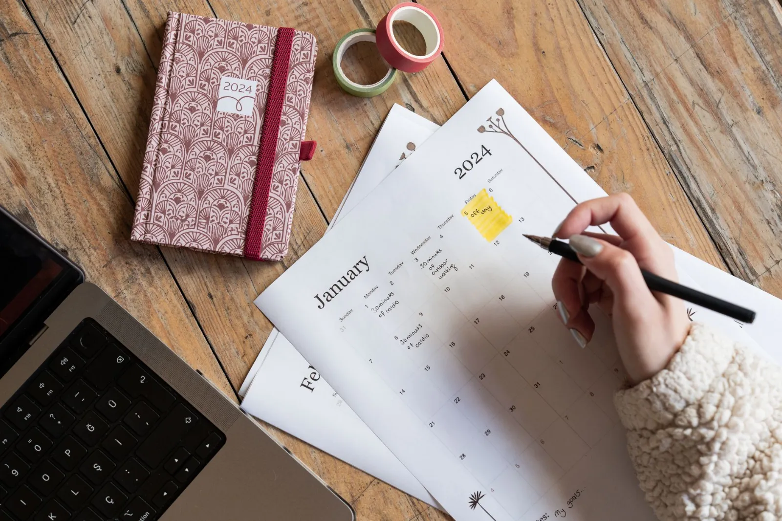 Hands writing in a planner next to a laptop