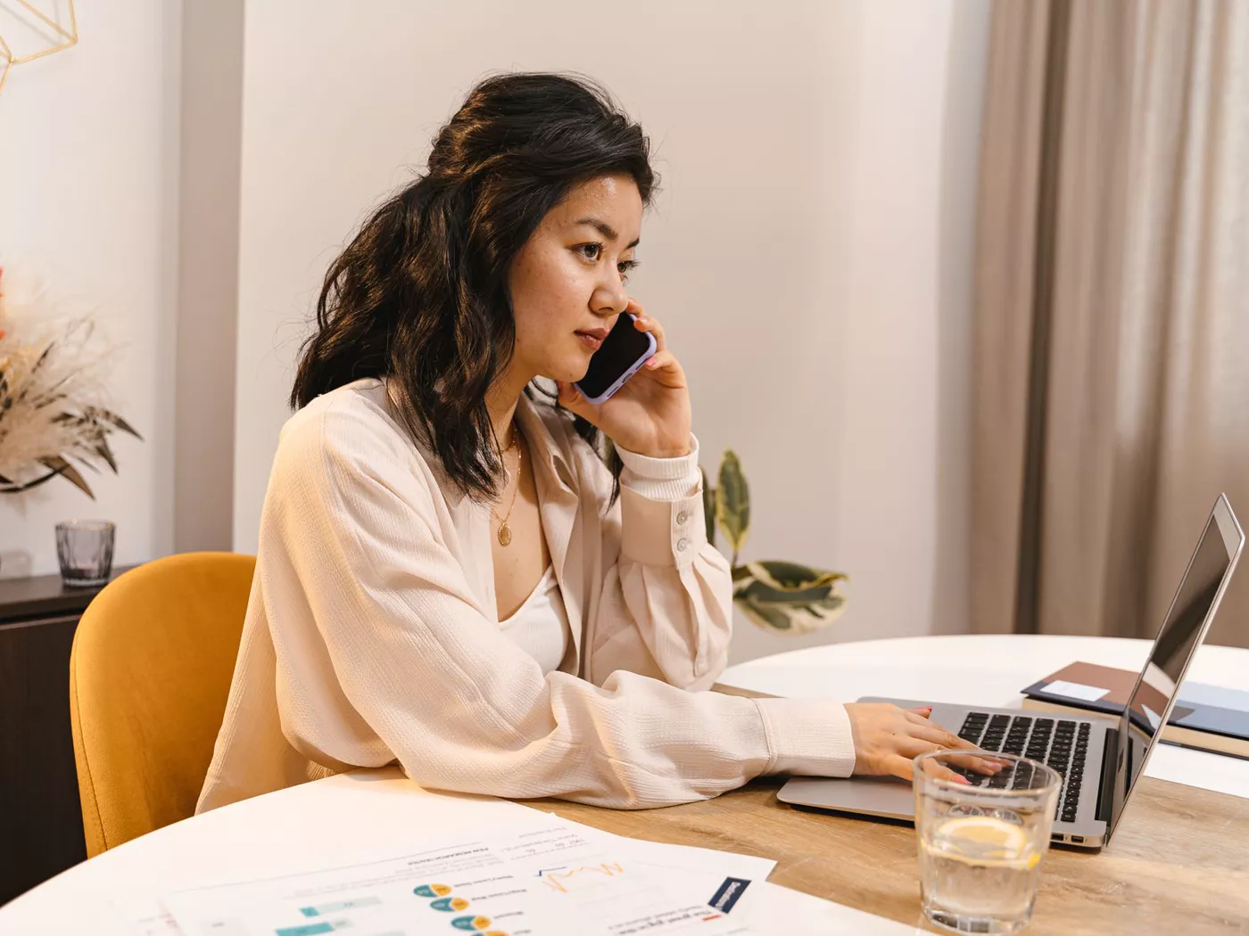 Adult woman working from home, using a laptop and mobile phone at a home office desk.